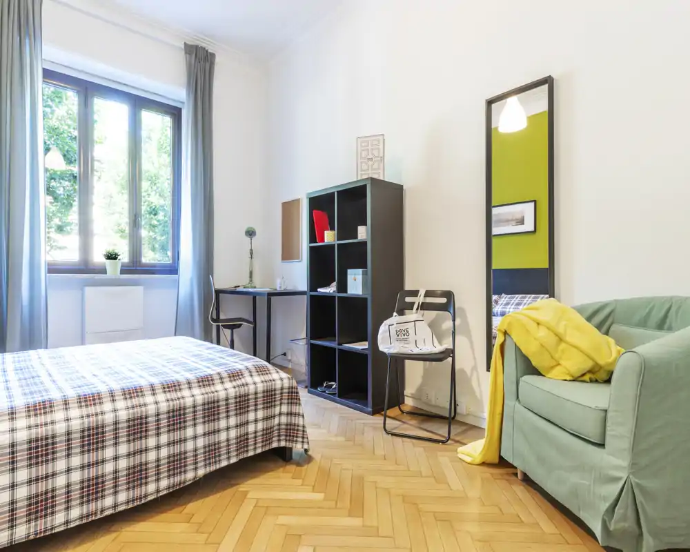 Dormitorio luminoso y bien presentado con una cama grande con funda a cuadros, un escritorio y estantería junto a la ventana y un sillón acogedor con una manta amarilla.