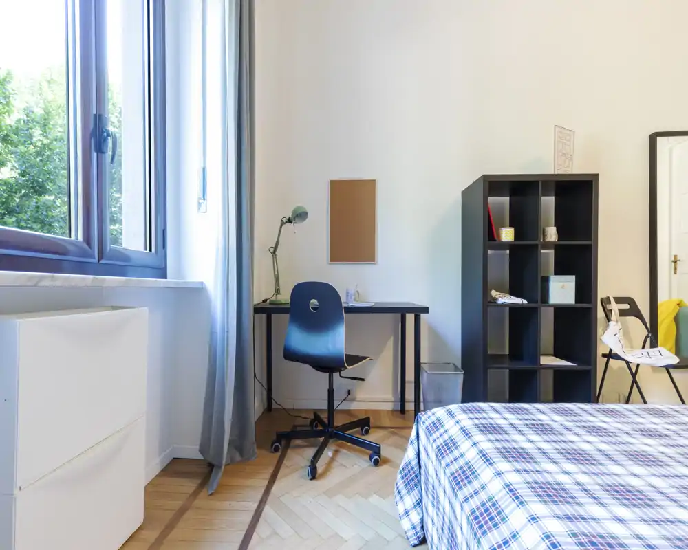 Dormitorio luminoso y ordenado con colcha a cuadros, ventana que deja entrar luz natural, escritorio y estantería para almacenamiento — ideal para estudiante o una persona.