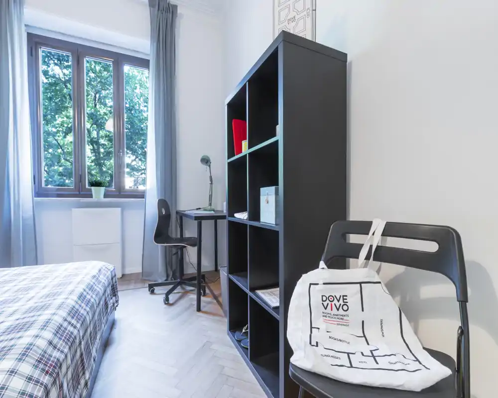 Dormitorio luminoso y bien decorado con cama de cuadros, escritorio junto a la ventana y estantería abierta para almacenamiento—perfecto para un estudiante o una persona.