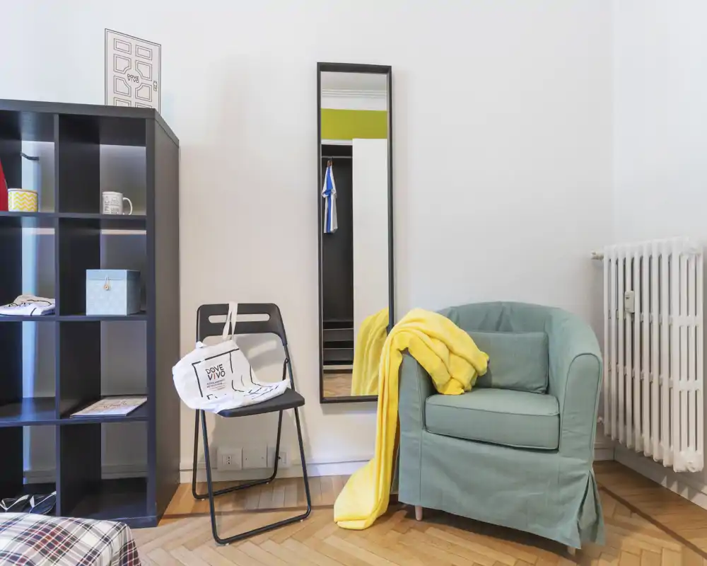 Rincón de salón acogedor con un sillón verde, una manta amarilla y un espejo de cuerpo entero. Las estanterías y el suelo de parquet aportan una sensación hogareña y luminosa, ideal para anuncios de alquiler.
