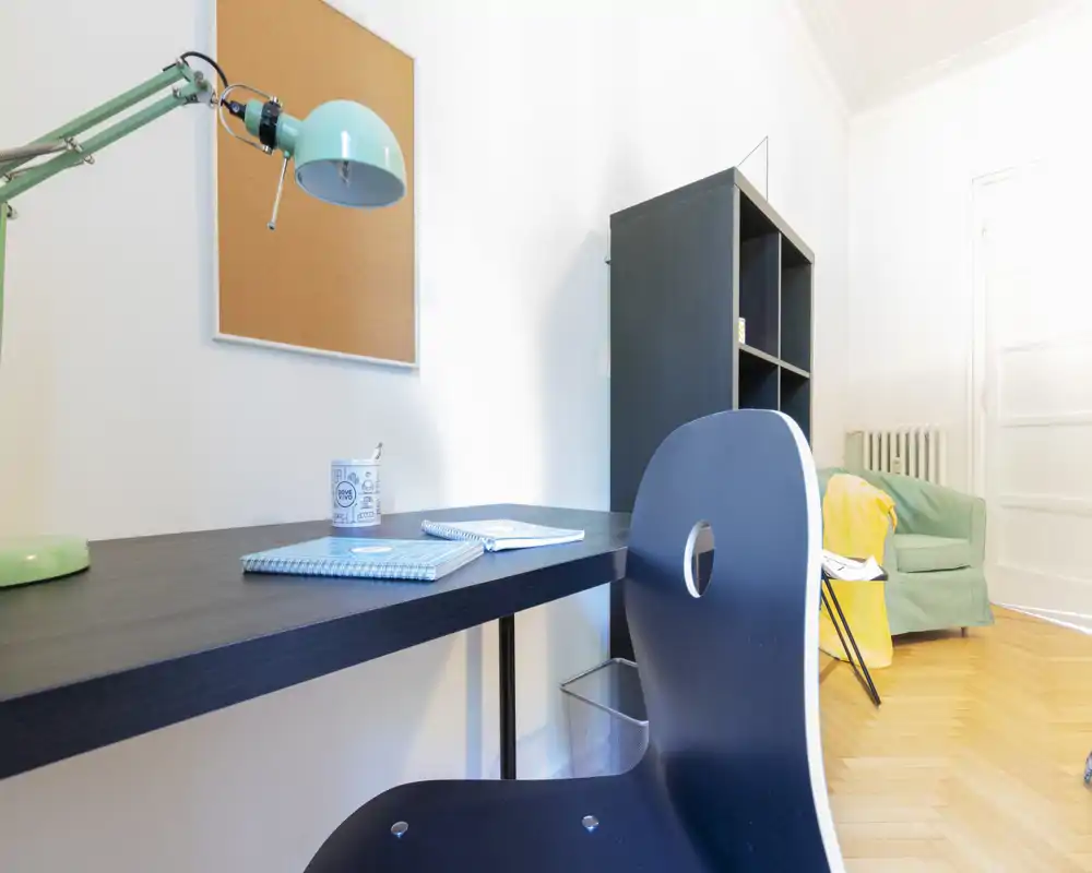 Zona de estar luminosa y cuidada con escritorio y un sillón cómodo — ideal para teletrabajo y descanso. Se aprecia una zona de trabajo con lámpara, estantería y asiento con luz natural.