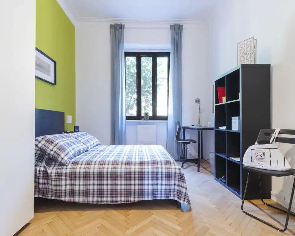 Dormitorio luminoso y ordenado con cama doble vestida con ropa de cuadros, escritorio junto a la ventana y estantería negra; suelo de parquet en espiga y cortinas suaves crean un ambiente acogedor.