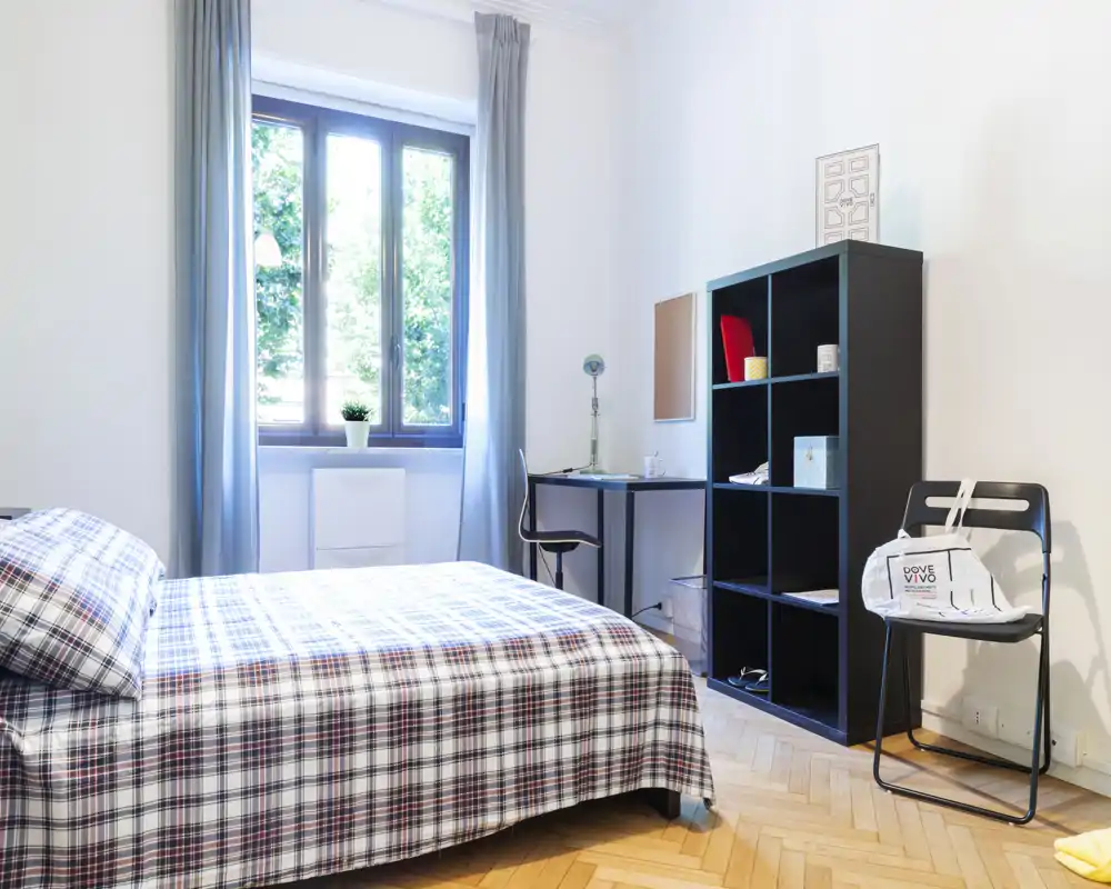 Dormitorio luminoso y ordenado con cama doble con colcha de cuadros, escritorio junto a la ventana y estantería para almacenamiento — ideal para una estancia cómoda.