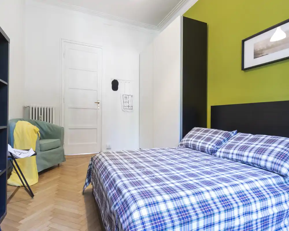 Dormitorio luminoso y bien decorado con cama doble de estampado a cuadros, pared de acento verde, armario y butaca acogedora; perfecto para anuncios de alquiler.
