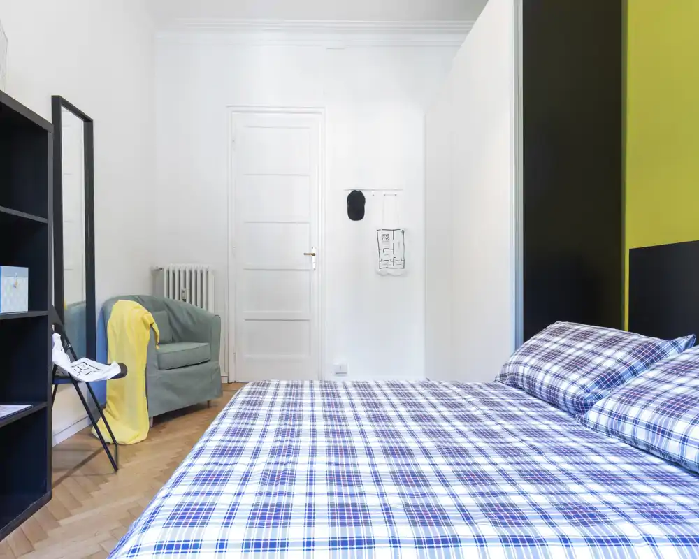 Dormitorio moderno y luminoso con una gran cama de cuadros en primer plano, un sillón acogedor y almacenamiento sencillo. Líneas limpias y paneles en contraste crean un espacio atractivo para un anuncio de alquiler.