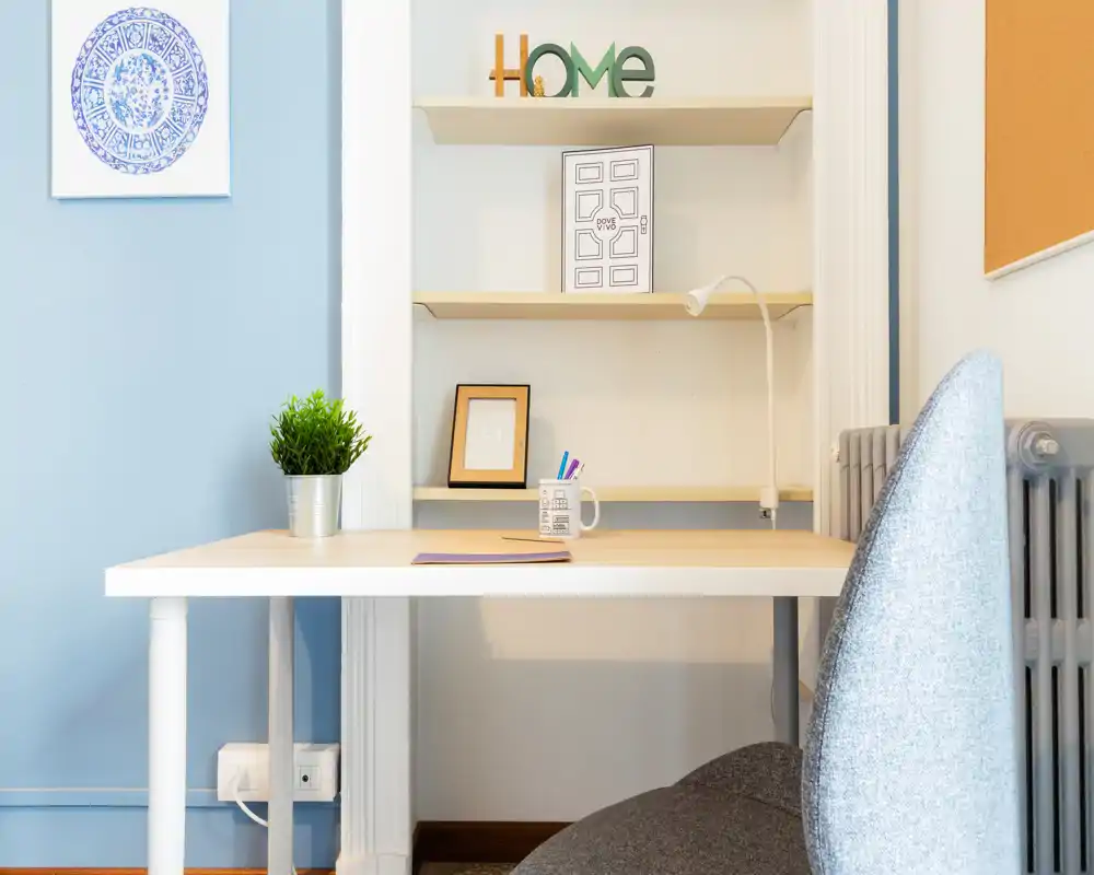 Rincón de oficina en casa luminoso y limpio con escritorio blanco, silla gris moderna, estantería y detalles decorativos como una planta y un marco.