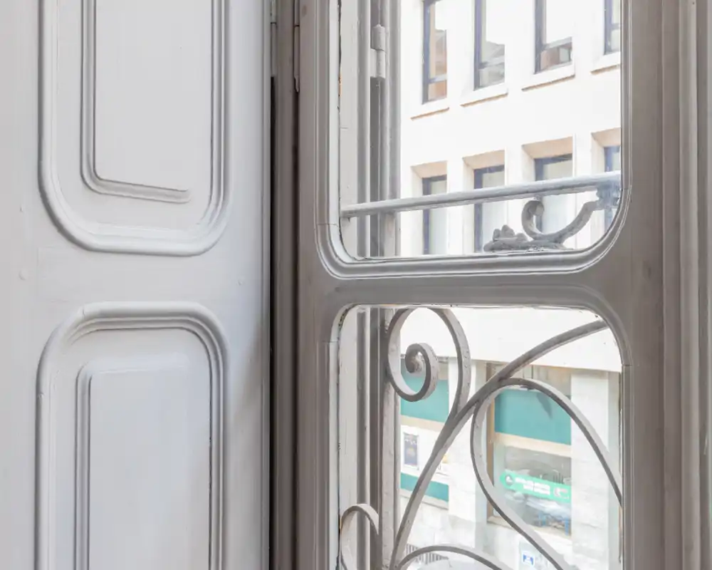 Vista en primer plano a través de una ventana decorativa y una barandilla de hierro forjado con vistas a la calle; luz natural suave y marco de madera pintada.