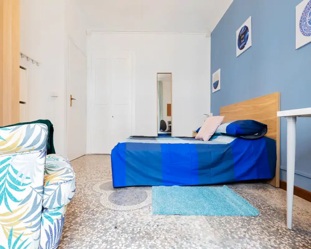 Dormitorio luminoso y moderno con cabecero de madera, ropa de cama azul y cojines decorativos. Suelo de mosaico, pequeña alfombra junto a la cama y un sillón cómodo en primer plano.