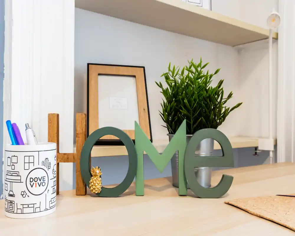 Plano cercano luminoso y cuidado de un rincón del salón: letras decorativas 'Home', planta en maceta y marco sobre una superficie de madera clara, perfecto para un anuncio acogedor.