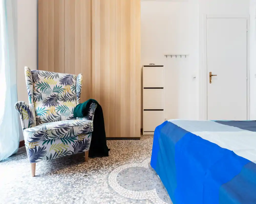 Rincón de dormitorio luminoso y bien presentado con un sillón estampado y una cama parcialmente visible con ropa de cama azul; la luz natural y el armario de madera cálida crean un ambiente acogedor.