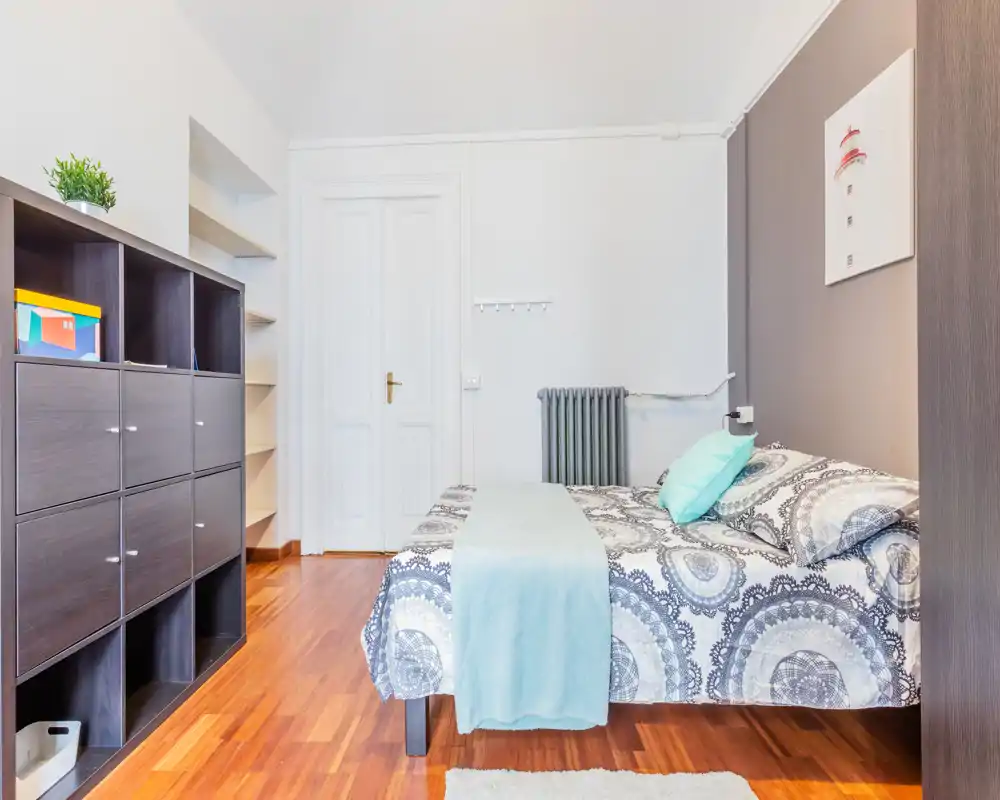 Chambre lumineuse et bien mise en scène avec un lit double habillé de linge à motifs, un jeté clair et des coussins. Parquet, étagères et accents doux pour une ambiance chaleureuse et moderne, parfaite pour une annonce.