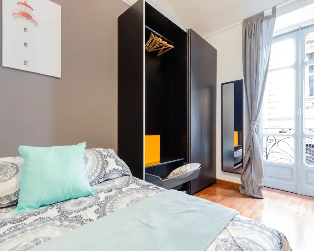 Chambre lumineuse et bien mise en scène avec linge de lit à motifs, coussins confortables et une grande fenêtre apportant une belle lumière naturelle.