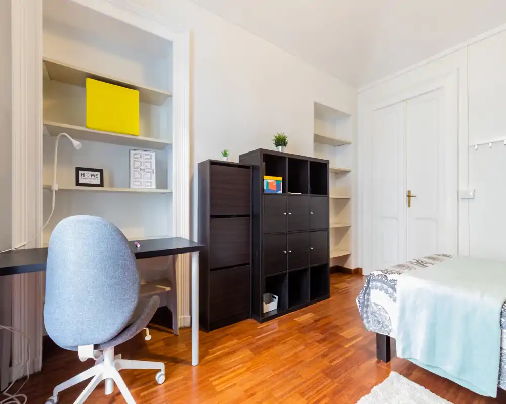 Chambre lumineuse et rangée avec lit simple, bureau et étagères ; parquet et textiles apportent une atmosphère chaleureuse et fonctionnelle, parfaite pour un étudiant ou une personne seule.