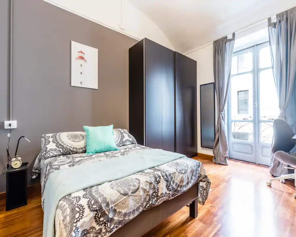 Chambre lumineuse et bien composée avec un lit double face à une grande fenêtre, armoire foncée et parquet chaleureux — confortable et moderne.