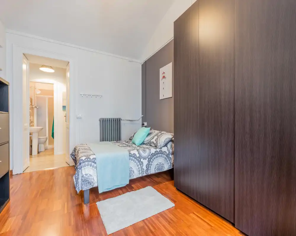 Chambre lumineuse et bien décorée avec un lit double habillé de linge à motifs, accents bleus et une grande armoire sombre. Sol en bois poli, petit tapis et salle de bains attenante visible par la porte.