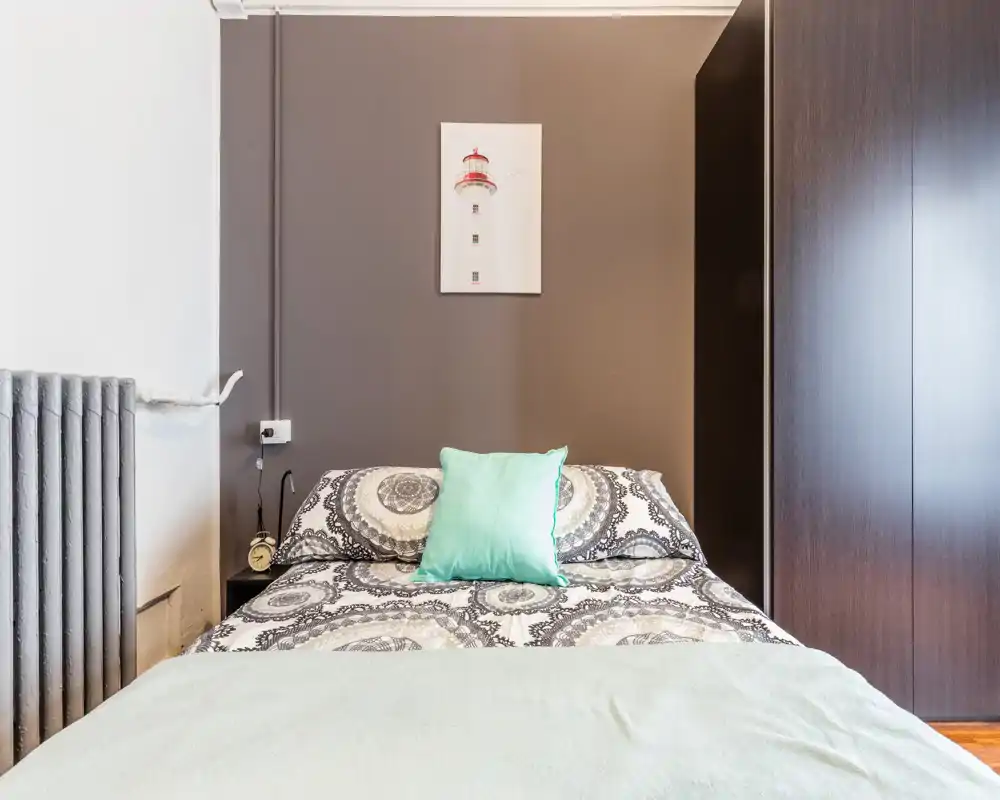 Chambre soignée avec un lit double centré, une literie à motifs et un coussin vert menthe; une armoire moderne et un éclairage doux créent un espace de repos chaleureux.