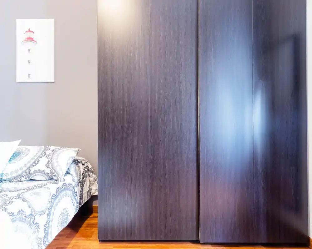 Coin de chambre lumineux avec un lit partiellement visible à gauche et une grande armoire en bois sombre à droite ; style simple et moderne avec un sol en bois chaleureux.
