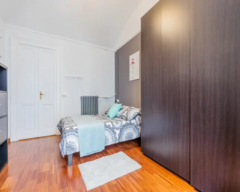 Chambre lumineuse et bien agencée avec un lit double, linge de lit à motifs et une grande armoire foncée; sol en bois chaleureux et touches douces pour une atmosphère accueillante.