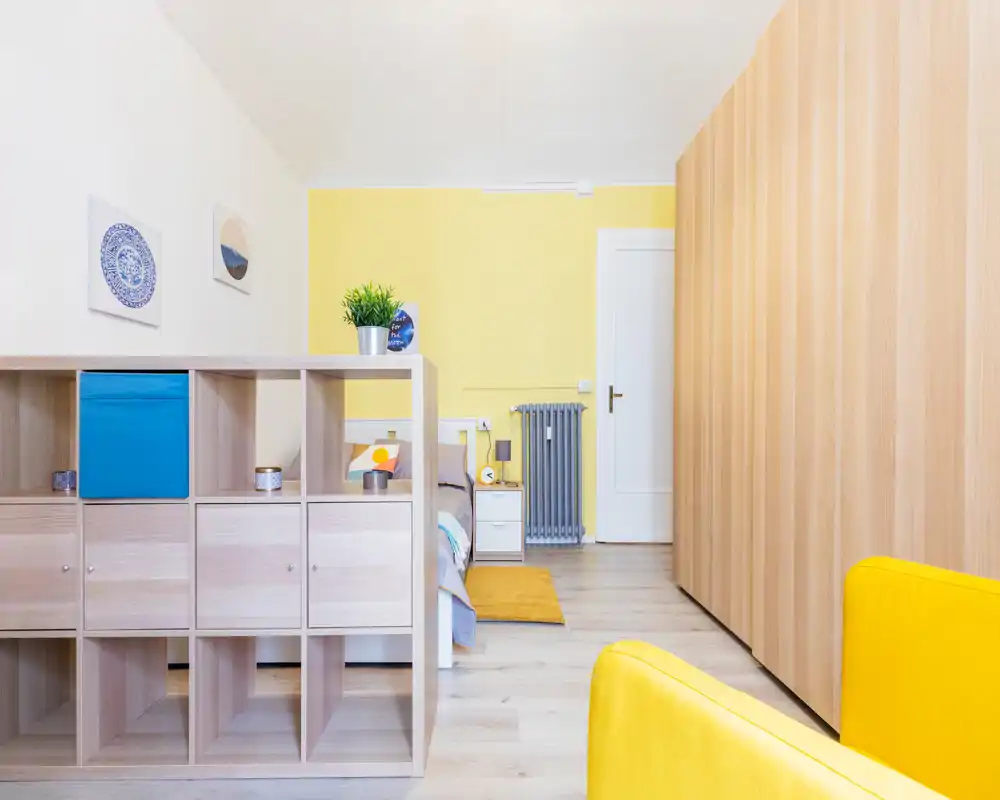 Chambre moderne et lumineuse aux accents jaunes, lit visible derrière une étagère basse, textiles assortis et rangements pour une atmosphère chaleureuse et soignée.