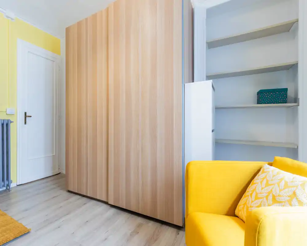 Espace de vie moderne et lumineux avec un fauteuil jaune vif, des étagères intégrées et une grande armoire en bois — lignes nettes et accents chaleureux pour un coin cosy.