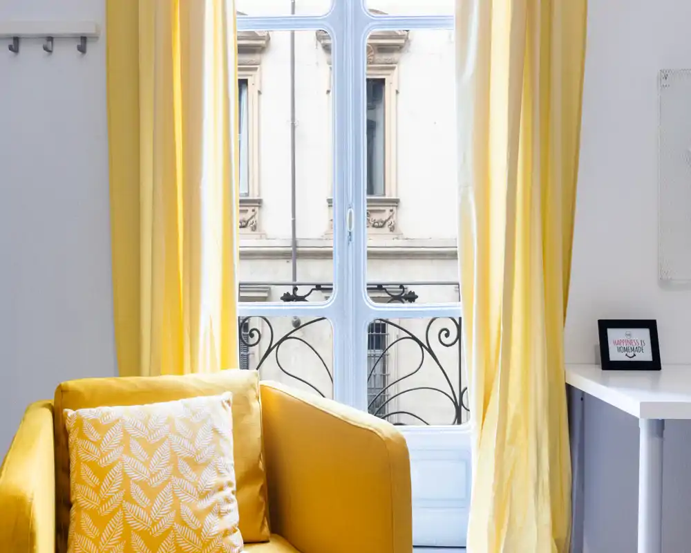 Espace séjour lumineux et élégant avec un fauteuil jaune et une grande fenêtre laissant entrer la lumière naturelle, rideaux assortis et coussin décoratif.