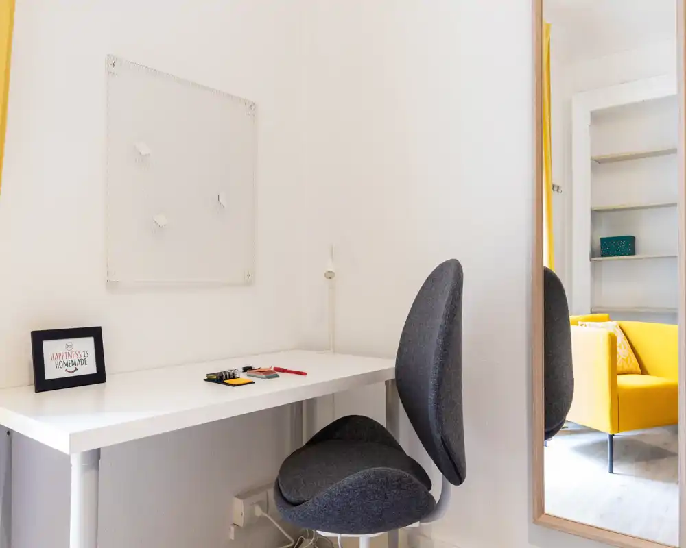 Coin séjour lumineux et compact avec un petit bureau et une chaise grise moderne ; un fauteuil jaune reflété dans le miroir apporte une touche vive.