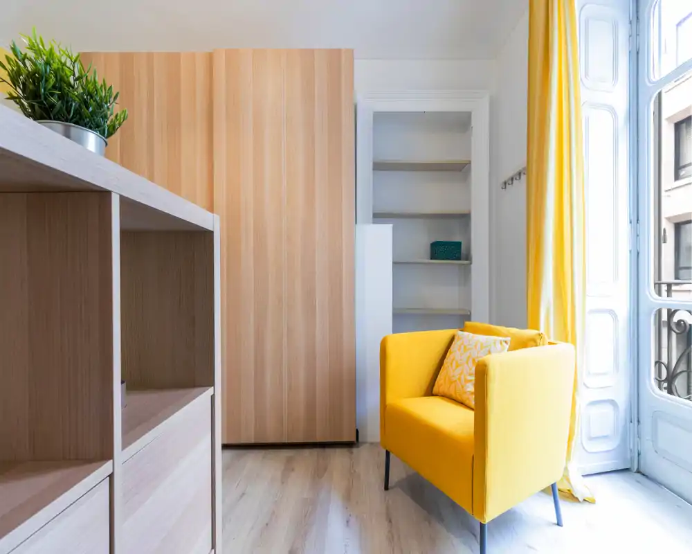 Coin salon lumineux et moderne avec un fauteuil jaune, rangements en bois clair et grande fenêtre ensoleillée offrant une ambiance fraîche et accueillante.