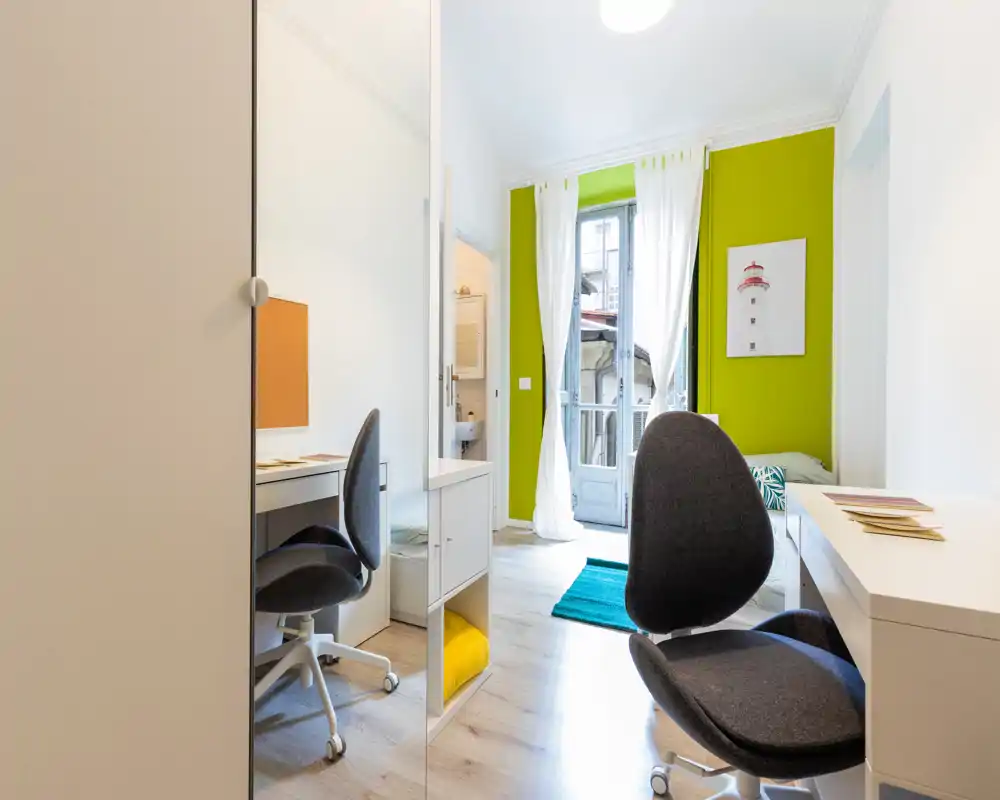 Chambre lumineuse et moderne avec coin bureau et accès au balcon. On note un mur vert, un lit partiellement visible, un bureau avec chaise ergonomique, un sol en bois clair et un tapis turquoise.