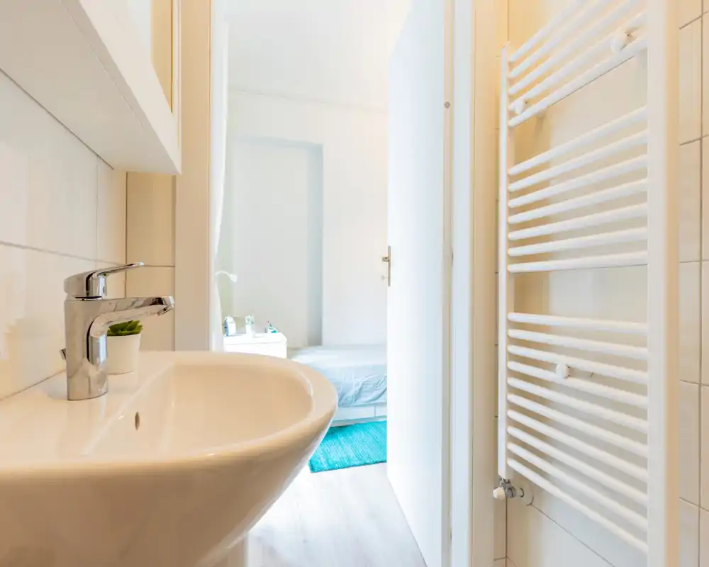 Salle de bain moderne et lumineuse avec lavabo en céramique blanche et radiateur sèche-serviettes mural; la porte donne sur une chambre visible en arrière-plan.