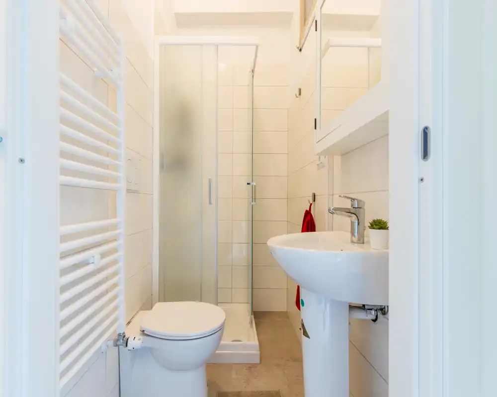 Salle de bain lumineuse et propre avec cabine de douche d'angle, lavabo sur colonne et WC ; carrelage moderne et sèche-serviettes.