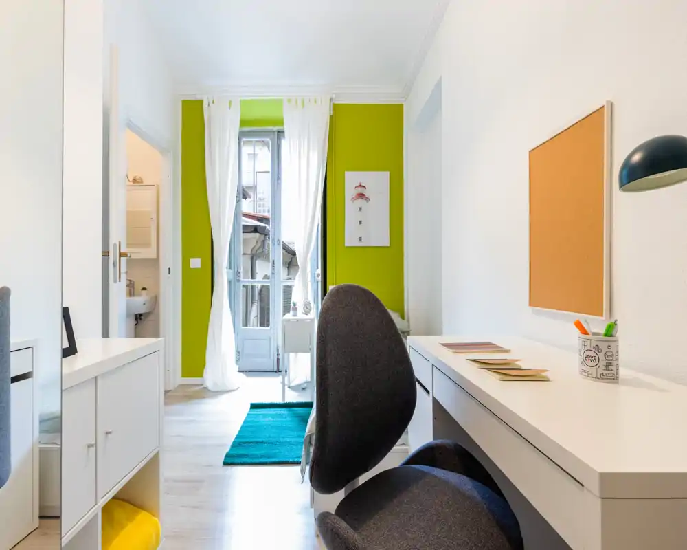Coin salon lumineux et moderne avec bureau, chaise ergonomique, tapis et accès au balcon. Meubles blancs et mur vert pour un espace de travail frais et aéré.