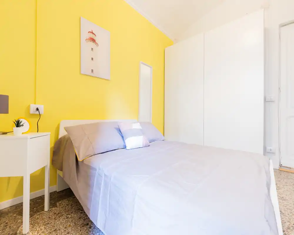 Dormitorio luminoso y bien presentado con cama doble, ropa de cama suave y una pared amarilla llamativa. Armario y mesita que completan un ambiente acogedor, perfecto para alquileres vacacionales.
