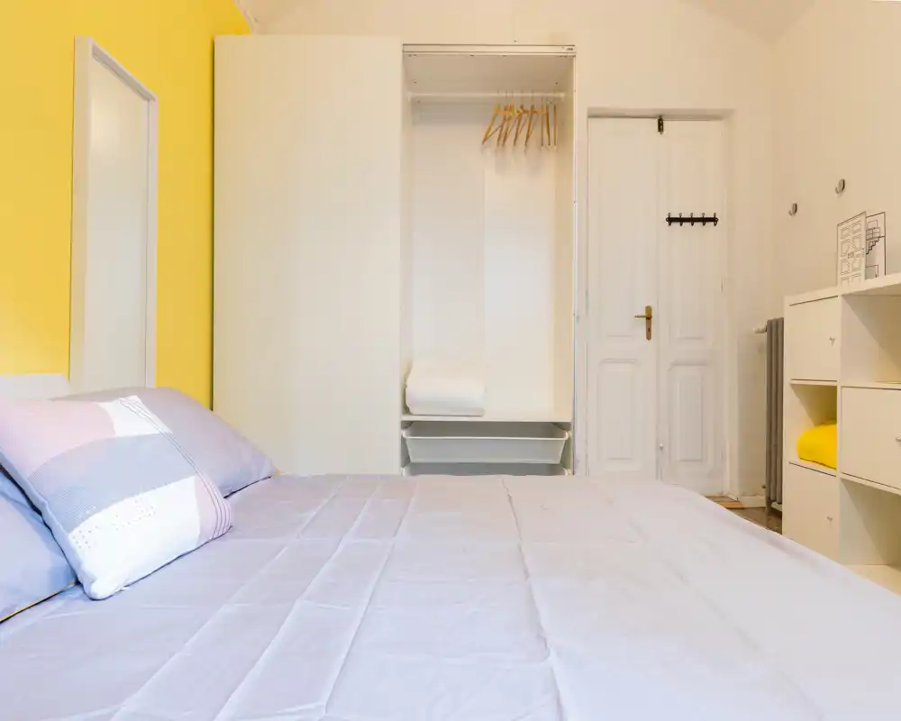 Dormitorio luminoso y ordenado con una cama grande en primer plano, un armario abierto empotrado y una puerta blanca; ropa de cama suave y una pared amarilla crean un ambiente fresco y acogedor.