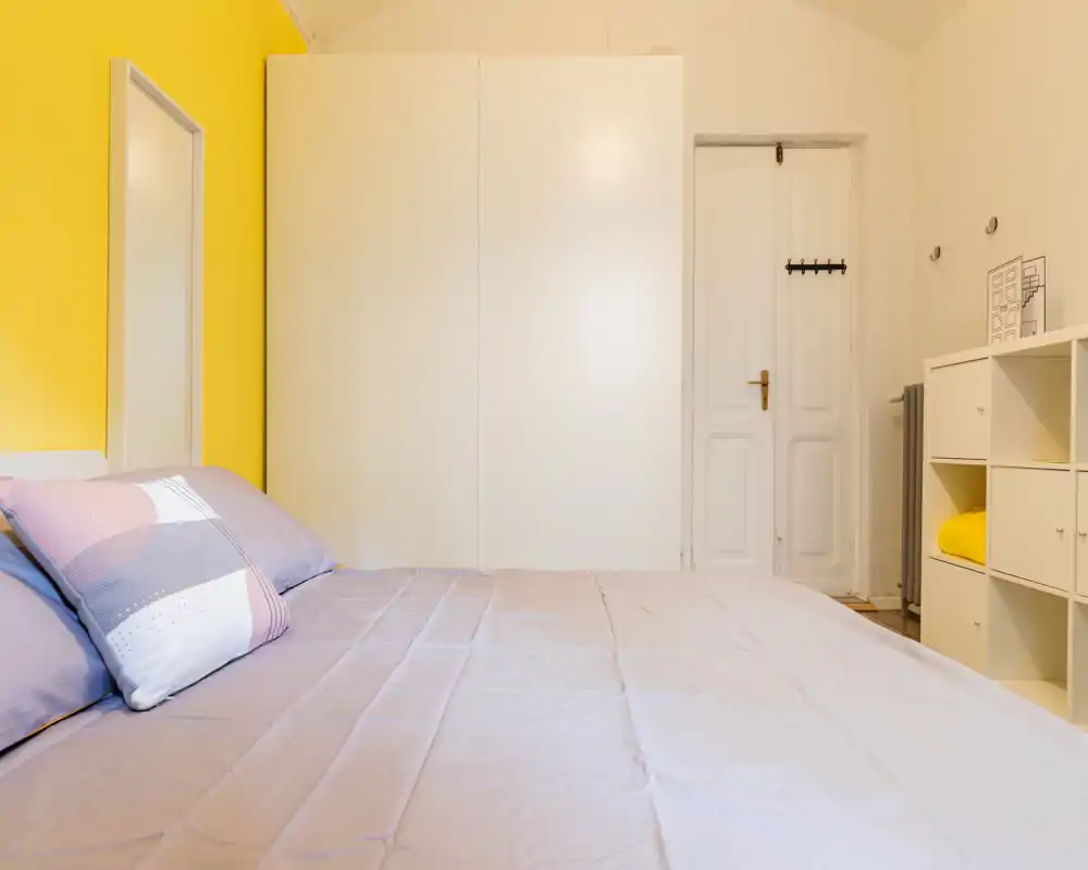 Dormitorio luminoso y ordenado con una cama amplia en primer plano, ropa de cama en tonos pastel y una pared amarilla como acento. Armario blanco y estantería modular ofrecen buen espacio de almacenamiento.