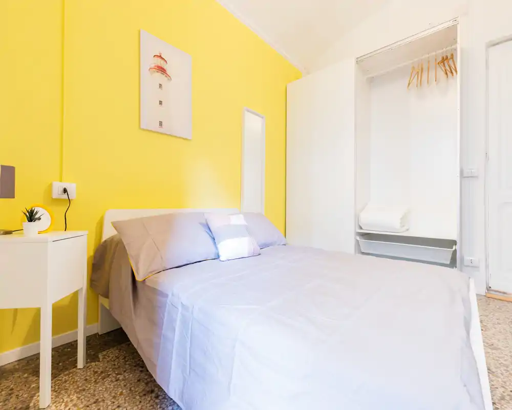 Dormitorio luminoso y ordenado con cama doble, ropa de cama gris y una alegre pared amarilla; armario empotrado abierto y una mesita completan el espacio.