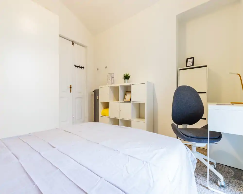 Dormitorio limpio y minimalista con una cama grande en primer plano, estantería blanca, escritorio y silla ergonómica — decoración luminosa y neutra, perfecta para alquileres.