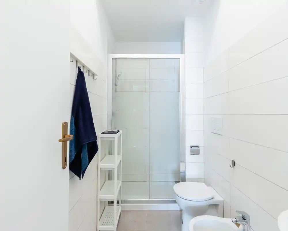 Salle de bain moderne et lumineuse avec douche à paroi vitrée, toilettes murales et carrelage blanc net ; étagère compacte et serviettes fraîches.