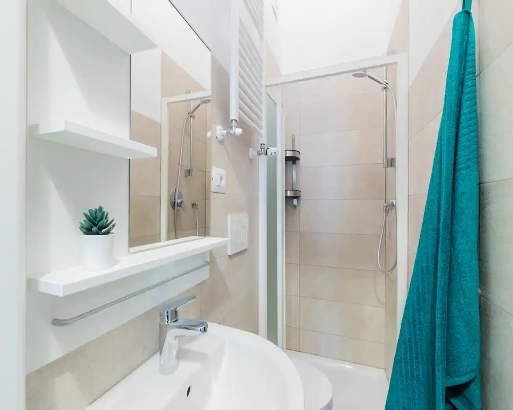 Salle de bain moderne et compacte avec douche d'angle, sanitaires muraux, lavabo blanc et serviette turquoise — lumineux et propre.