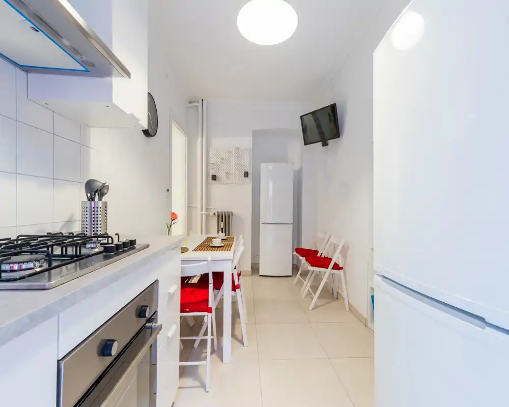 Cuisine moderne et lumineuse avec plaque à gaz, four et réfrigérateur. Petite table à manger avec chaises aux coussins rouges pour un coin repas accueillant.