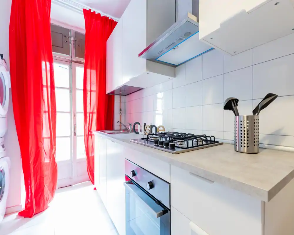 Cuisine moderne et lumineuse avec plaque à gaz, four encastré et rideaux rouges du sol au plafond laissant entrer beaucoup de lumière ; meubles blancs et crédence carrelée pour une ambiance fraîche et contemporaine.