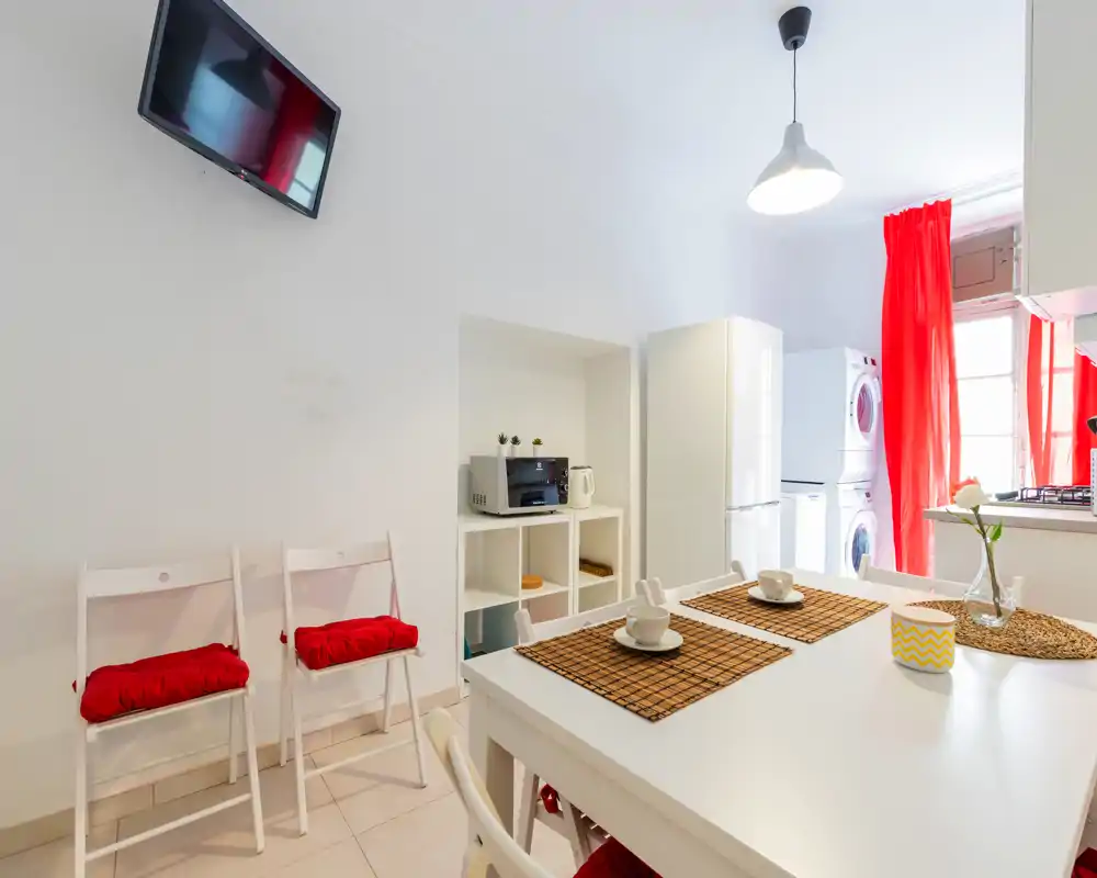 Cuisine compacte et lumineuse avec table blanche, lave-linge et sèche-linge empilés, réfrigérateur et rideaux rouges pour une ambiance moderne et fraîche.