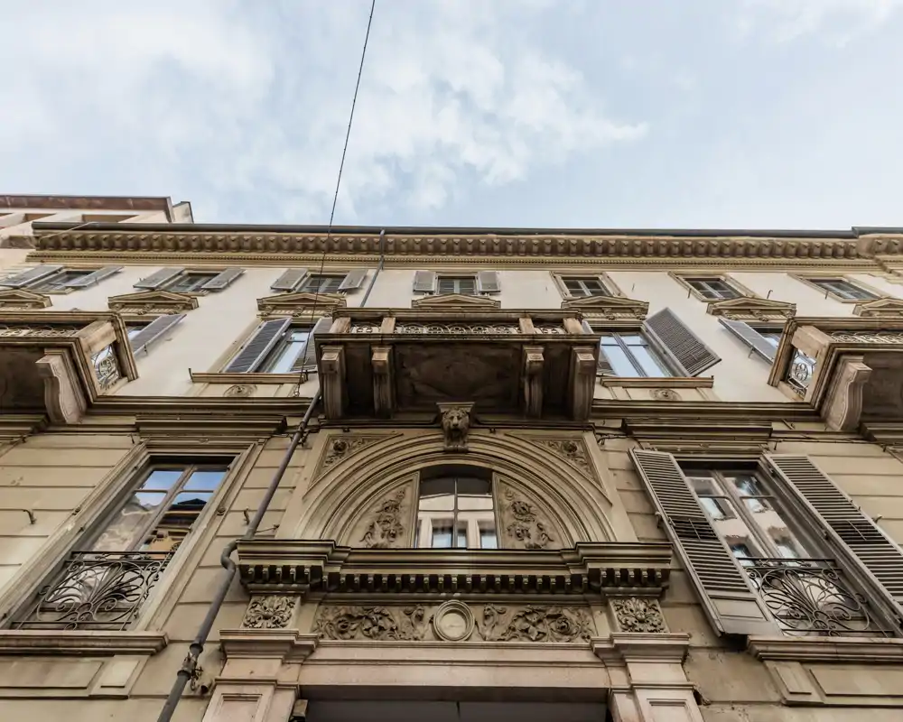 Façade extérieure d'un immeuble historique vue depuis la rue, présentant des ornementations en pierre, des balcons et des volets sous un ciel partiellement nuageux.