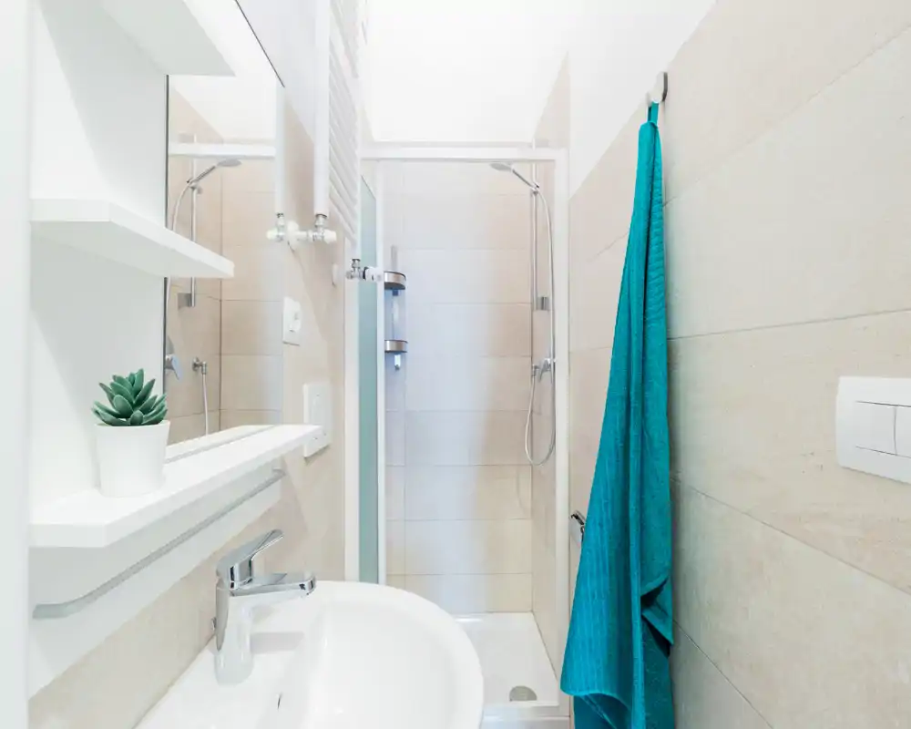 Salle de bains moderne et lumineuse avec douche d'angle, lavabo et miroir ; carrelage clair et serviette turquoise apportent une touche fraîche.