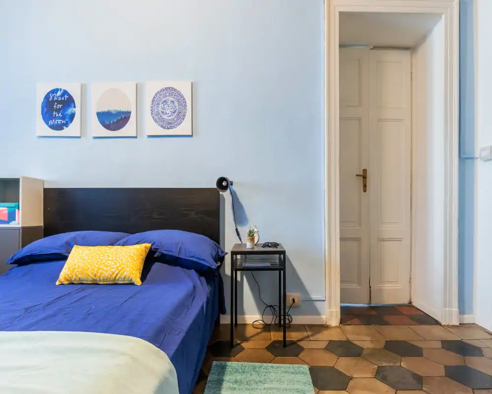 Camera da letto luminosa e ordinata con copriletto blu, cuscino giallo decorativo e comodino minimalista — linee pulite e accenti caldi perfetti per annunci.