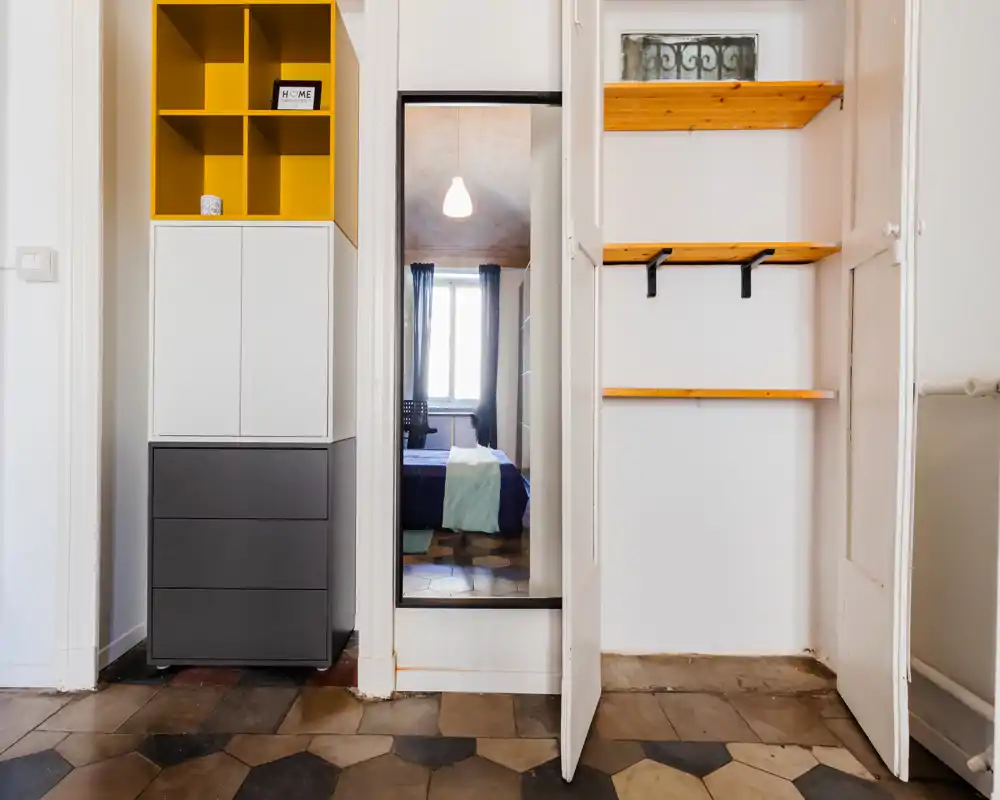Ingresso/corridoio con scaffalature e un armadietto alto; uno specchio a tutta altezza riflette una camera da letto con letto e finestra al centro.