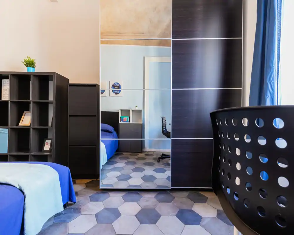 Camera da letto moderna con letto blu, armadio con ante a specchio e pavimento a piastrelle geometriche che creano uno spazio luminoso e di stile.