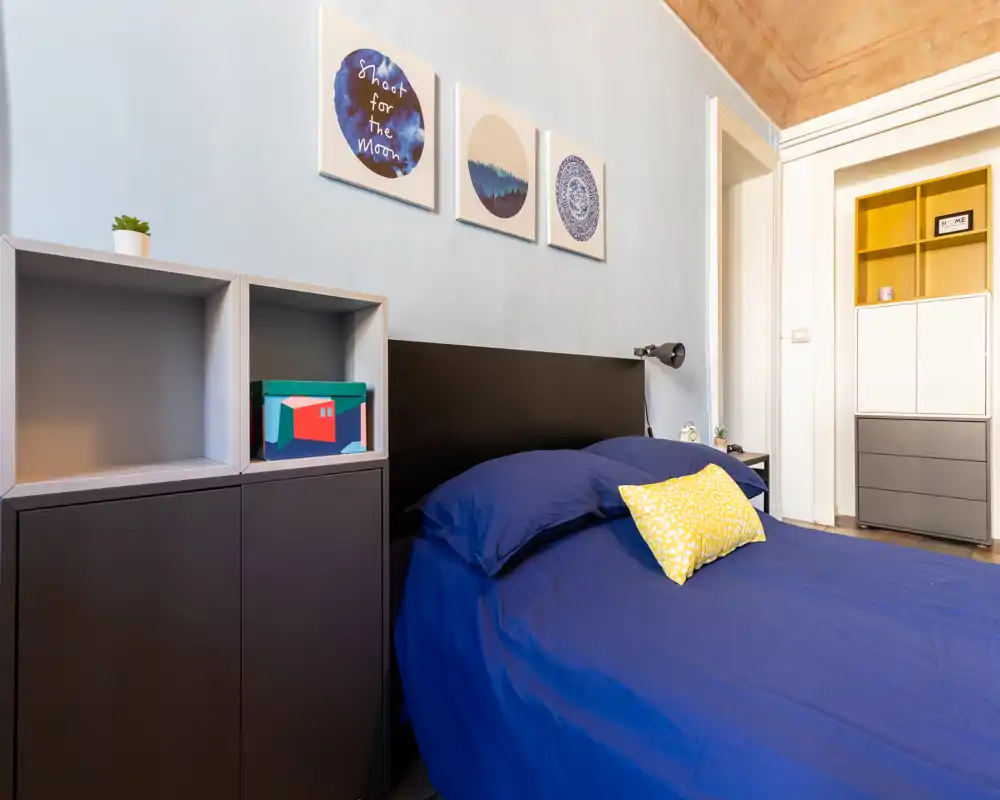 Camera da letto moderna e luminosa con copriletto blu navy, cuscino giallo a contrasto e scaffalature minimal. Quadri a parete e dettagli caldi sul soffitto aggiungono carattere mantenendo un’atmosfera pulita e ben illuminata.