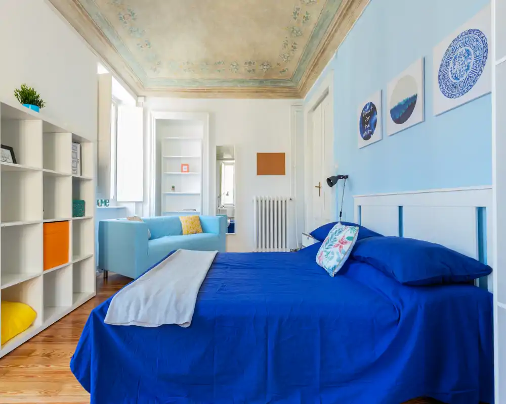Camera da letto luminosa e raffinata con un ampio letto blu in primo piano, cuscini coordinati e un divano azzurro vicino alla finestra. Soffitto decorato, libreria e molta luce naturale la rendono perfetta per un annuncio accogliente.