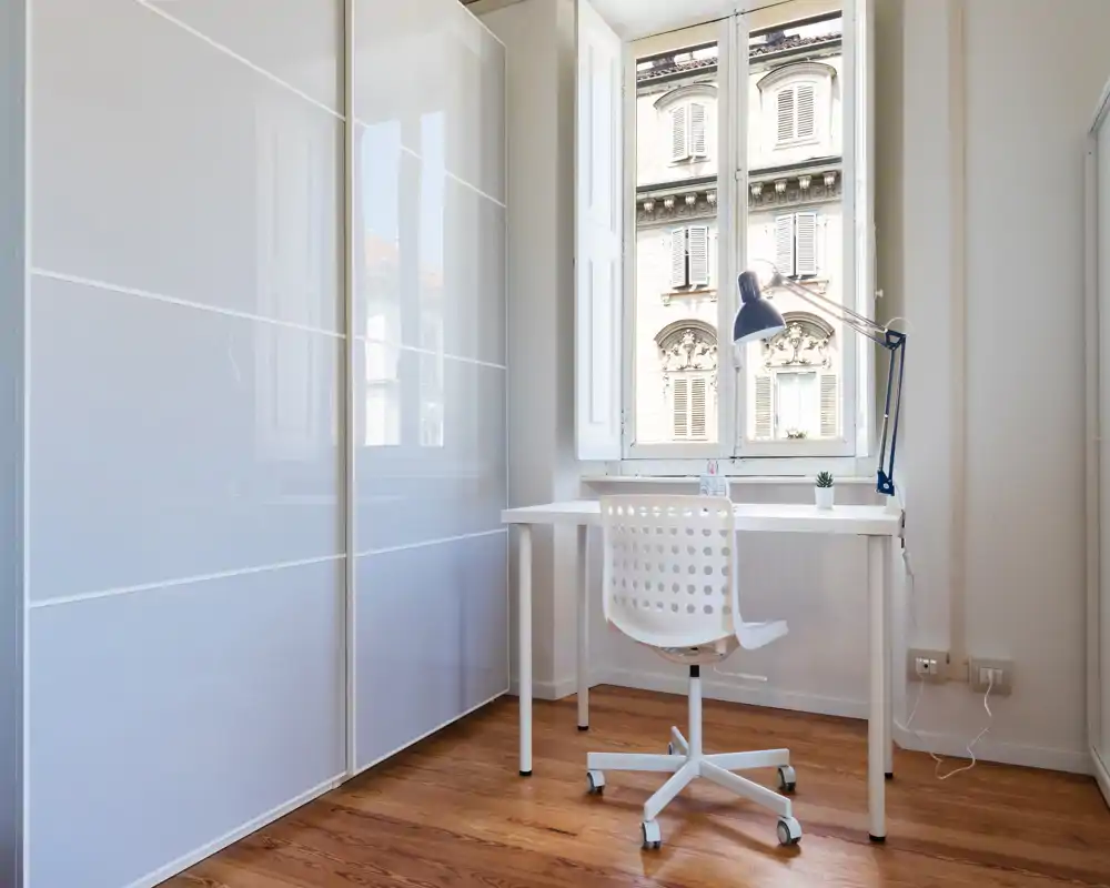 Angolo lavoro luminoso vicino alla finestra con scrivania bianca, sedia girevole e un ampio armadio lucido — perfetto per un ufficio domestico compatto.