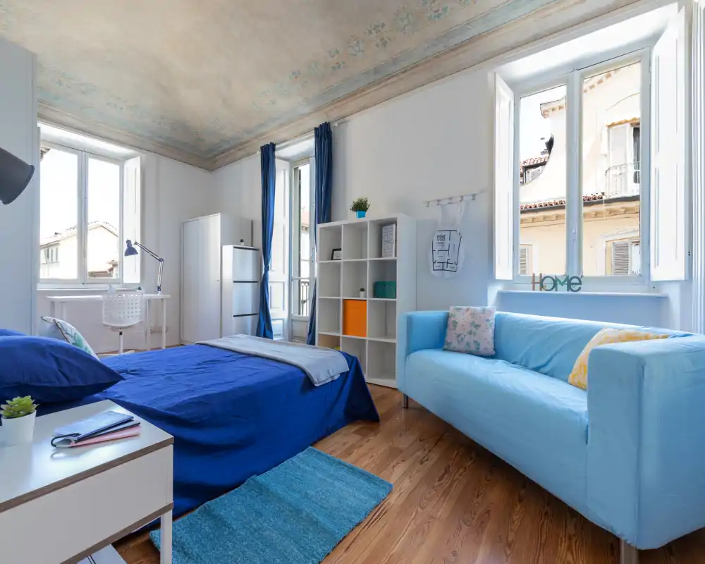 Camera da letto luminosa e ariosa con letto completo con biancheria blu, divano azzurro, pavimento in legno e grandi finestre che lasciano entrare molta luce naturale.